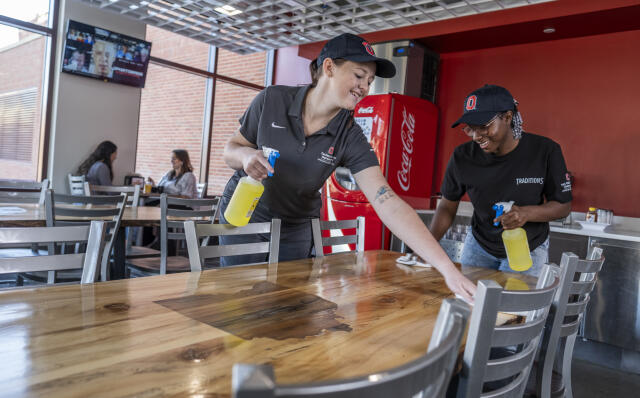 Two student employees clean a dining room table with cleaning spray and towels.