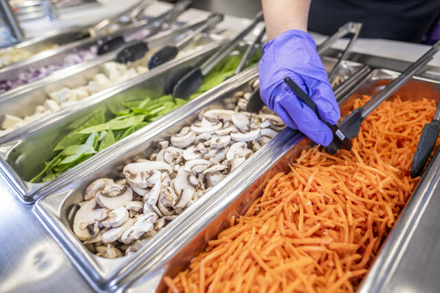 A staff member takes the temperature of shredded carrots on an ingredient bar.
