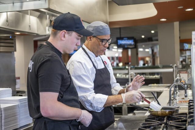 a chef teaches a student employee how to cook an omelet in a pan on a range.