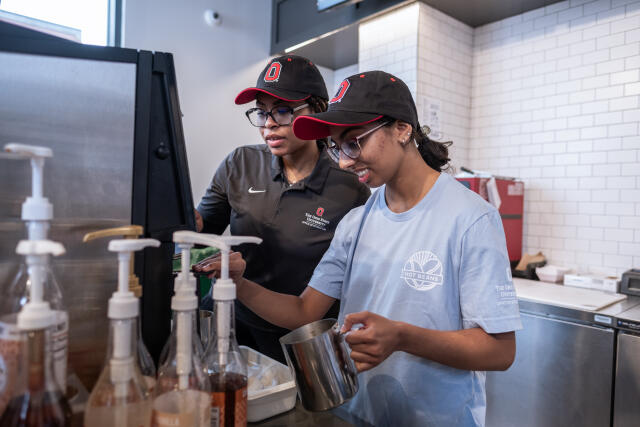 A student manager teaches a student employee how to steam milk on an espresso machine.